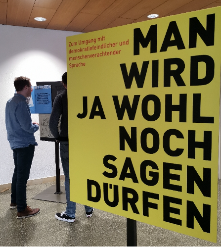 Im Vordergrund ist ein eine gelbe Tafel der Ausstellung zu sehen, auf der steht "Man wird ja wohl noch sagen dürfen". Im Hintergrund sind Besucher der Wanderausstellung zu sehen.